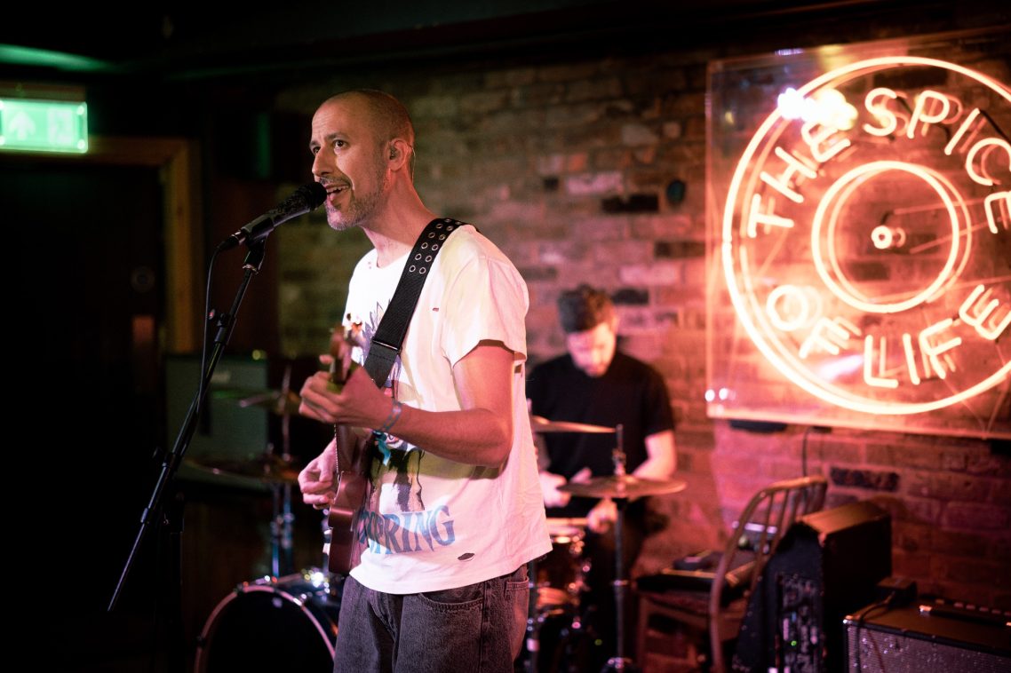 A musician sings and plays guitar on stage, with a drummer in the background.