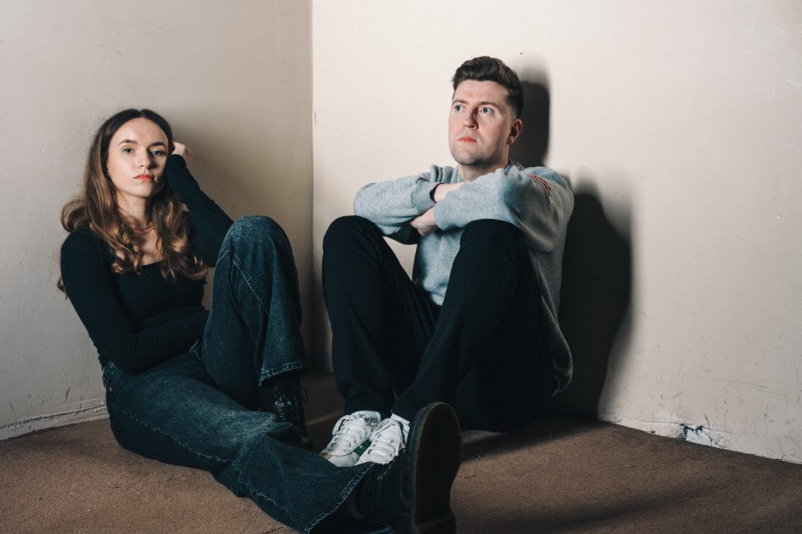 A woman and man sit against a wall, looking contemplative in a sparse room.