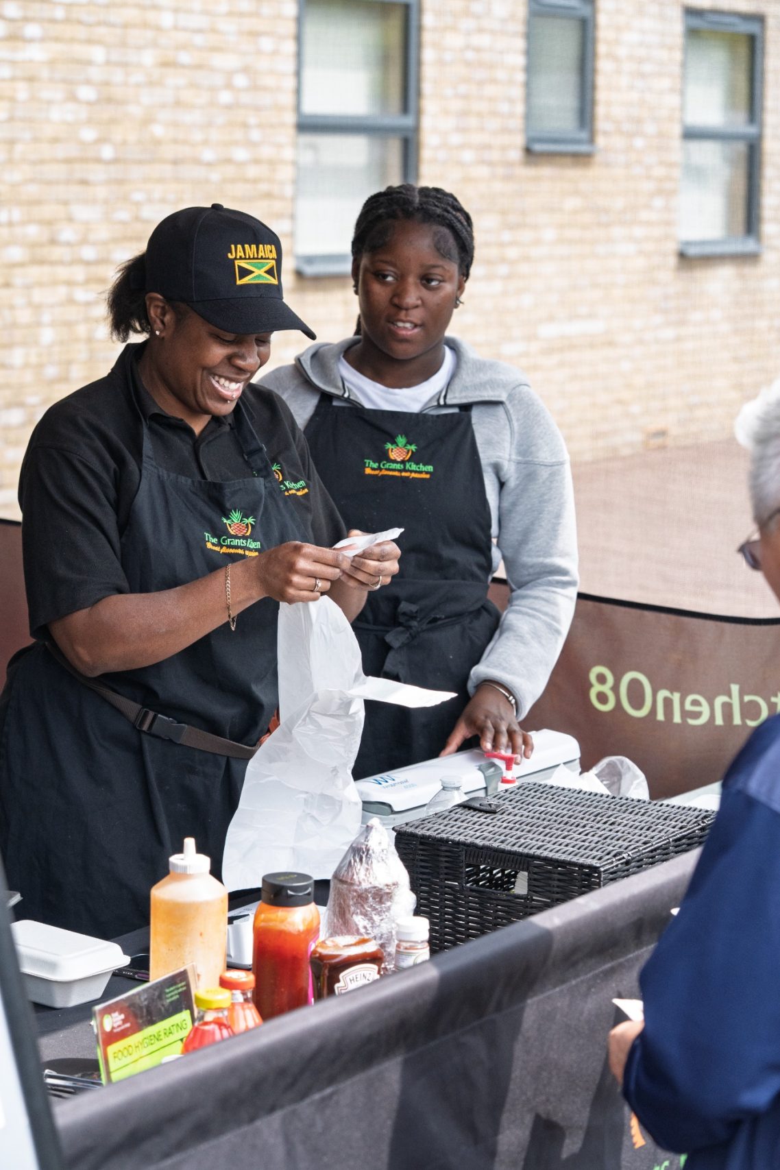 Two vendors at a food stall preparing orders, smiling and interacting with a customer.