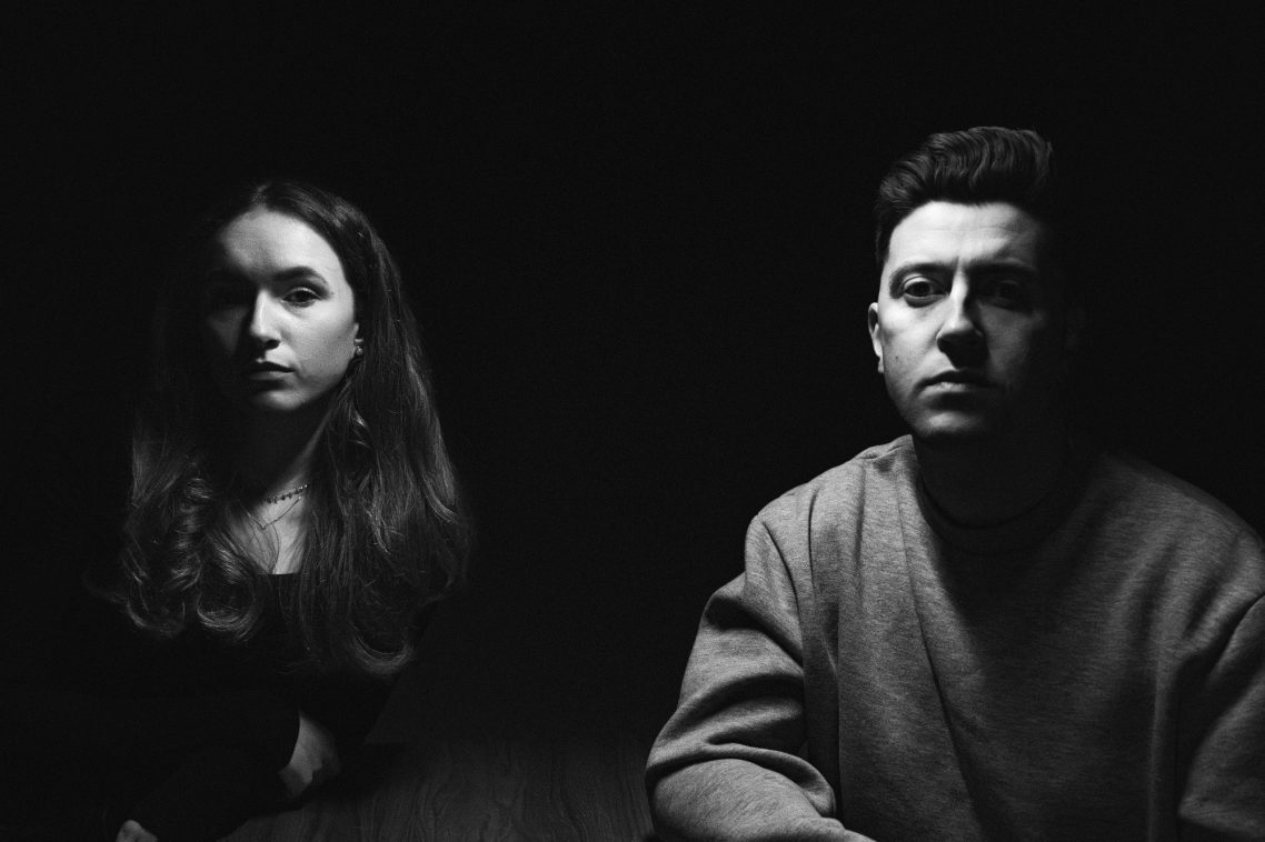 A man and a woman sit at a table in dim lighting, looking serious and contemplative.