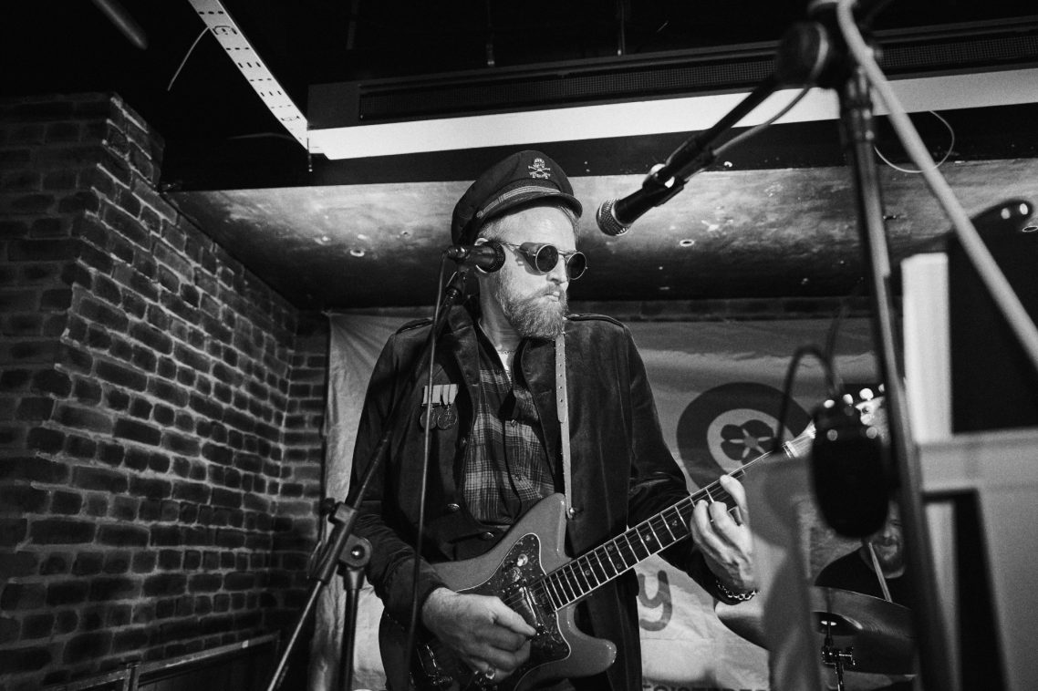 A musician playing guitar, wearing sunglasses and a hat, in a dimly lit venue.
