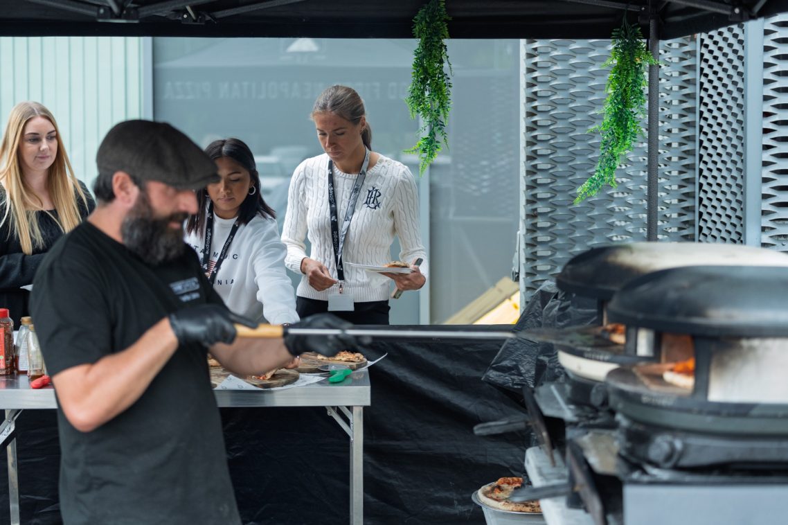 A chef using a pizza oven while three onlookers observe nearby.