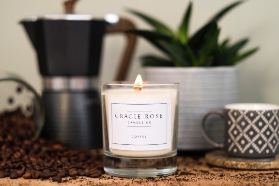 Coffee beans surround a lit candle, a plant, and a coffee maker on a countertop.