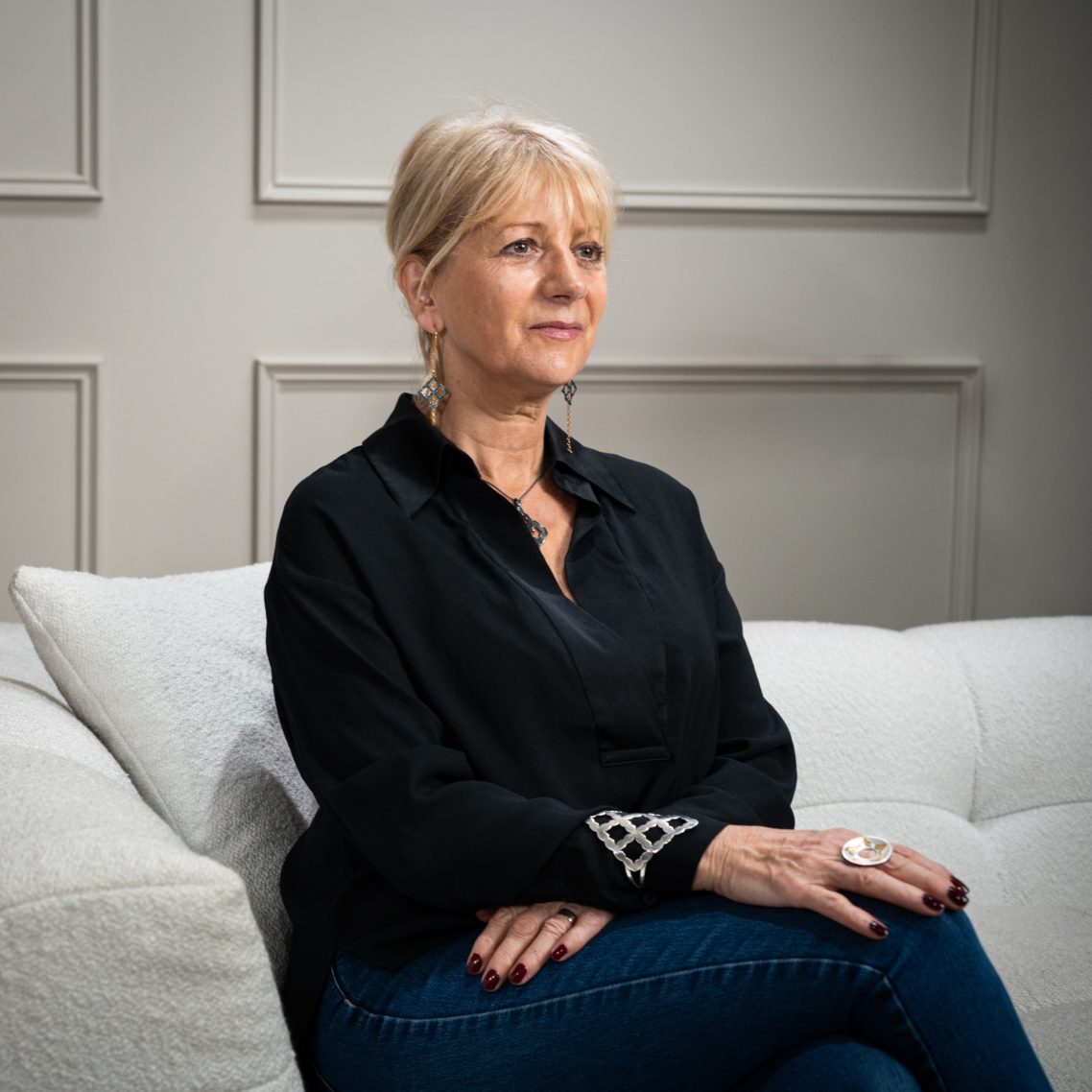 A woman with shoulder-length blonde hair sits on a sofa, wearing a black shirt and jeans.