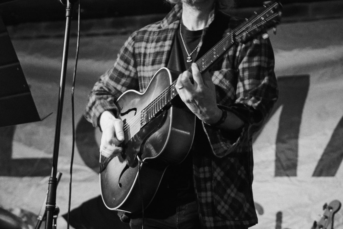 Person playing an acoustic guitar on stage, dressed in a checkered shirt.