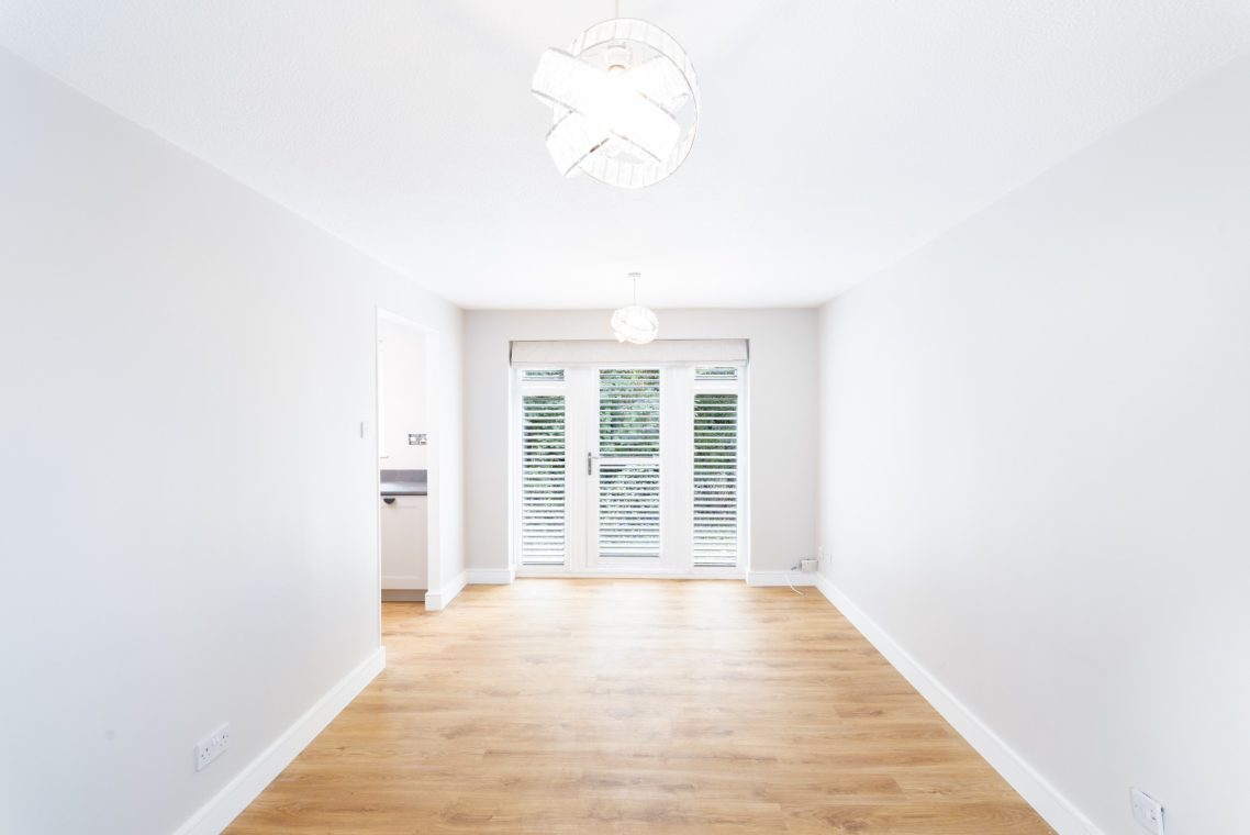 Bright, empty room with wooden floor and large windows leading to a garden outside.