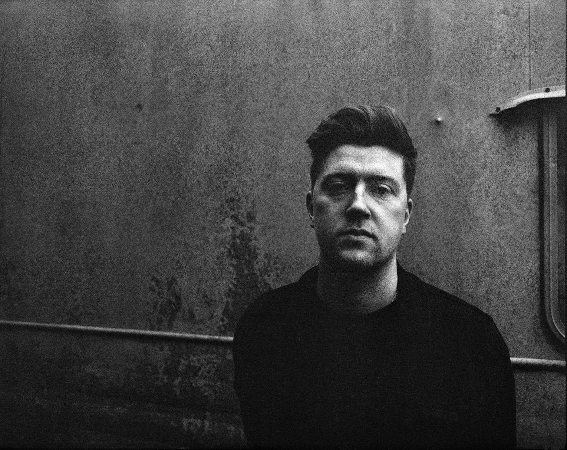 A man with short hair and glasses, looking intently, against a textured background.