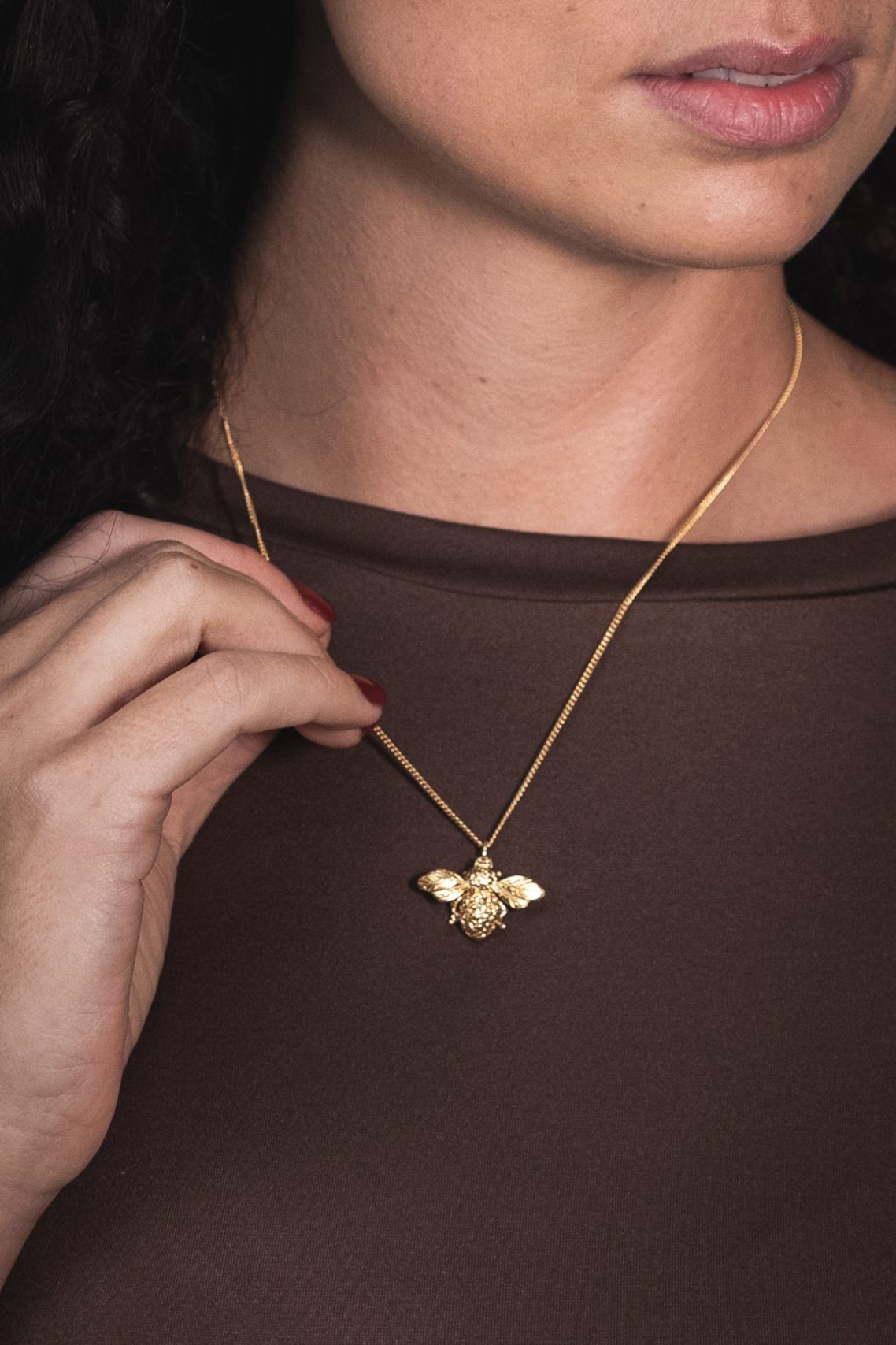 A close-up of a woman holding a gold bee pendant necklace, wearing a brown top.