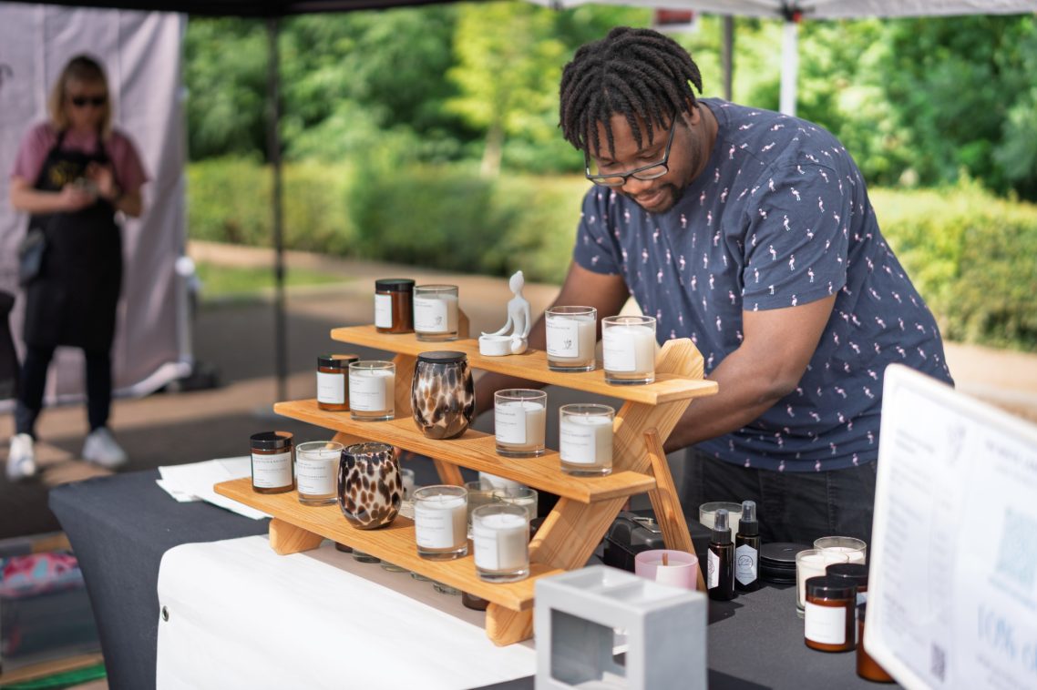 A man arranges scented candles on a wooden display at an outdoor market.
