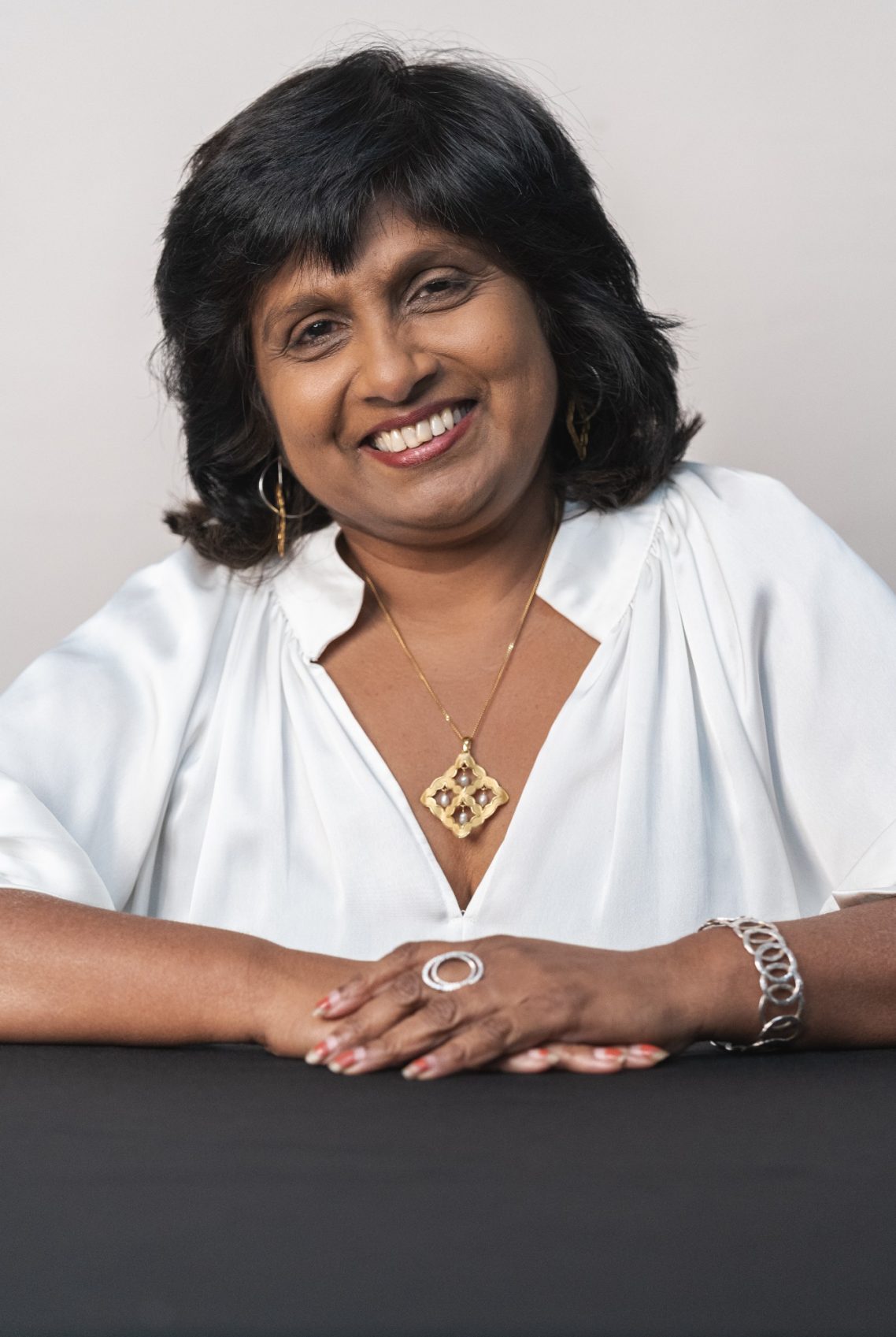 Smiling woman with short black hair, wearing a white top and jewellery, seated at a table.