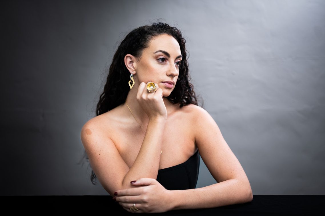 A woman with long curly hair poses thoughtfully in a black top, wearing statement earrings.
