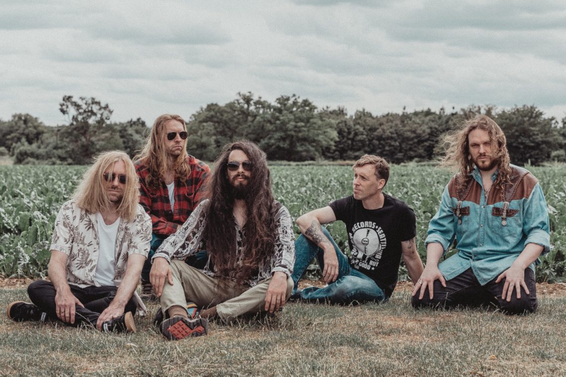 A five-member band seated in a grassy field, surrounded by trees under a cloudy sky.