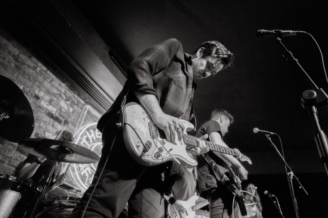 Band performing on stage, with a guitarist in focus against a brick backdrop.