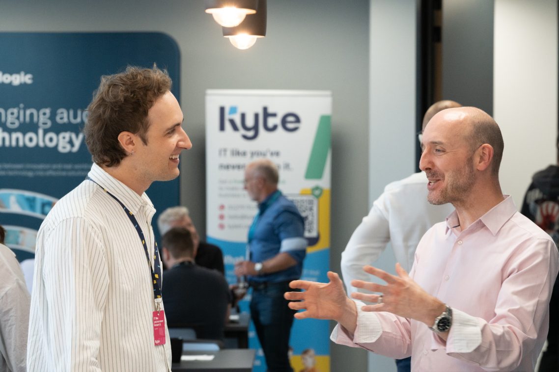 Two men engaged in conversation at a technology event, with banners in the background.
