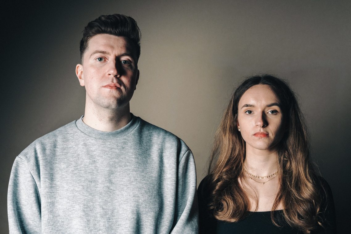 A man and woman standing side by side against a dark background, both looking directly at the camera.