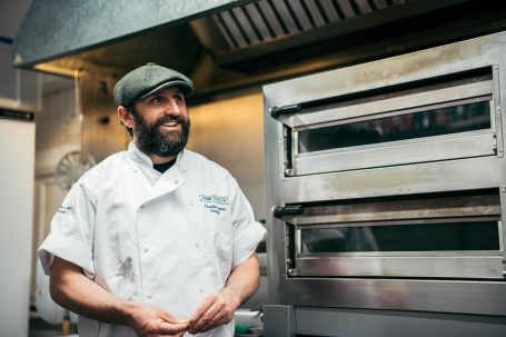 A smiling chef in a kitchen, wearing a cap and white uniform, stands near an oven.