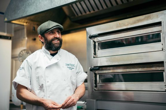 A smiling chef in a kitchen, wearing a cap and white uniform, stands near an oven.