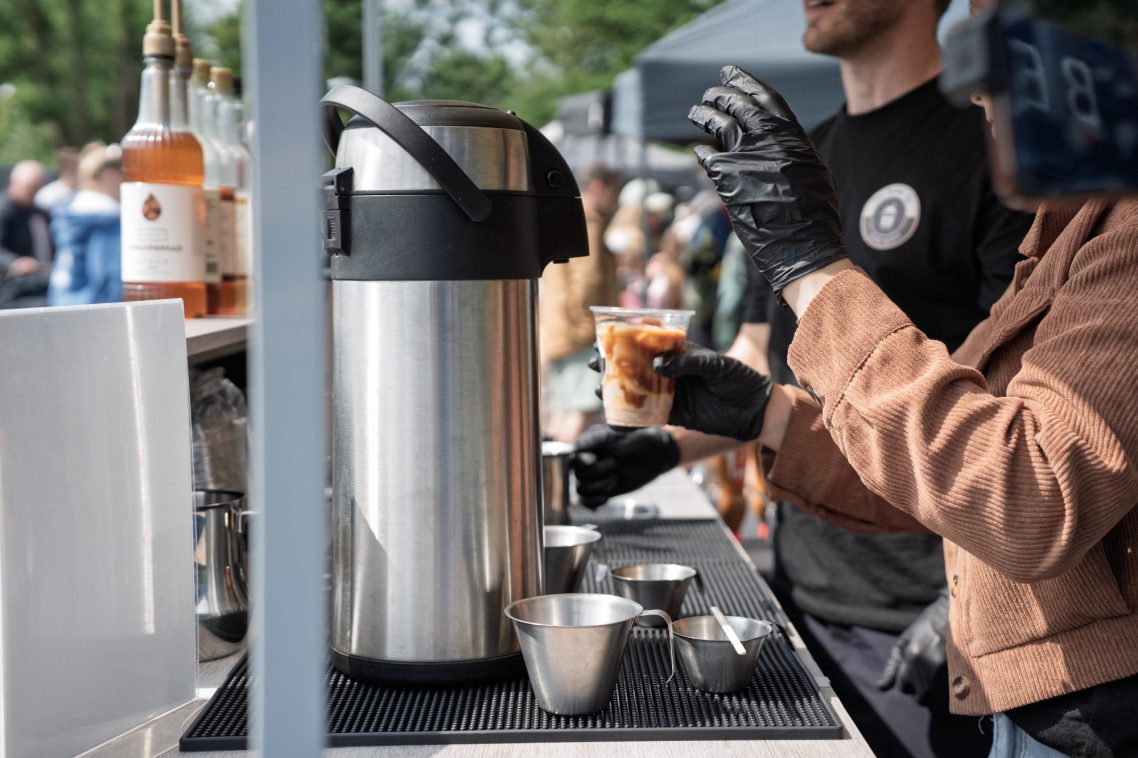 Barista serving coffee from a silver thermos at a busy outdoor event.