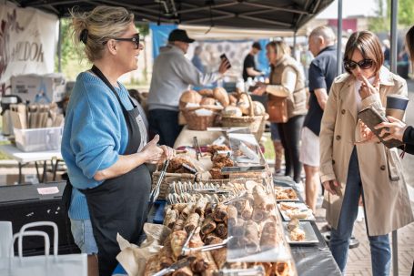 A market stall with baked goods, a vendor serving customers, and shoppers browsing.
