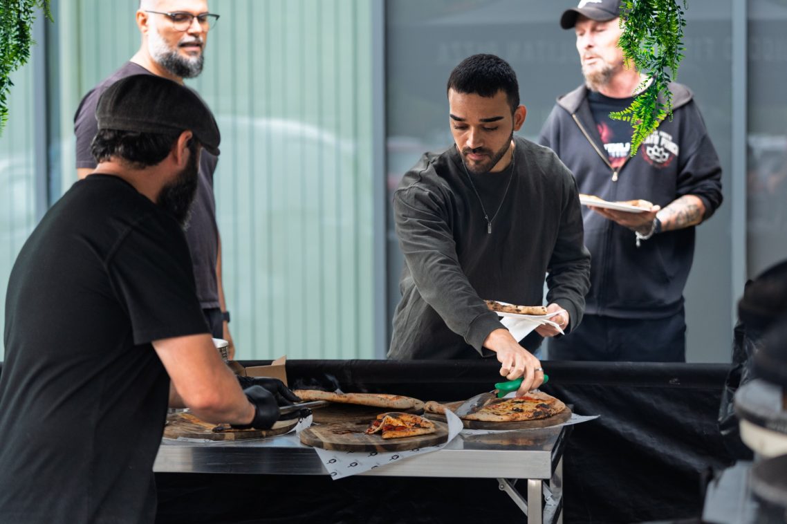 Two men serving food at an outdoor gathering, with another person in the background.