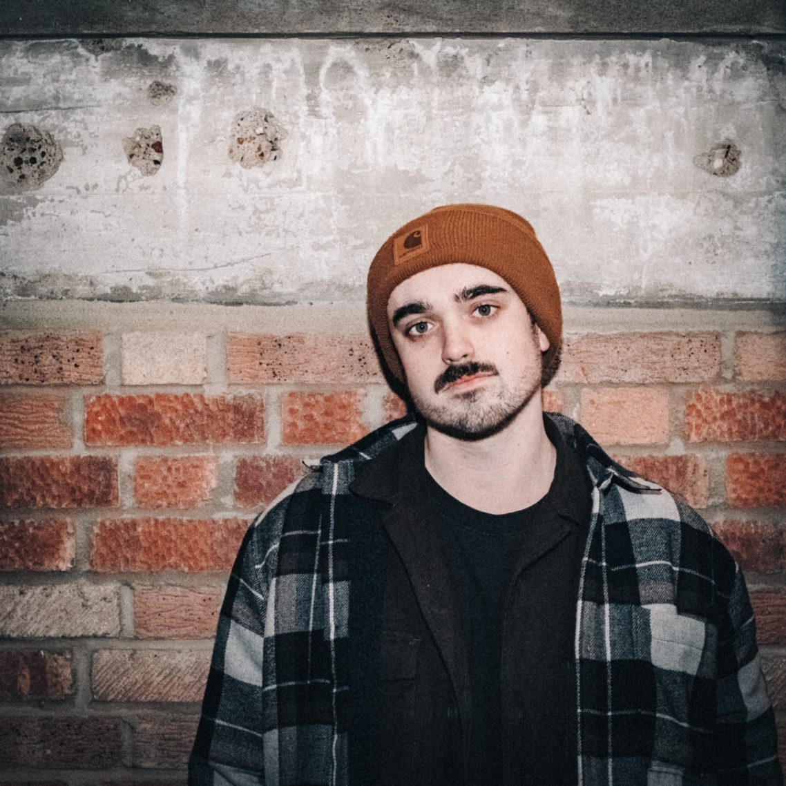 Young man in a brown beanie and plaid shirt standing against a brick wall.