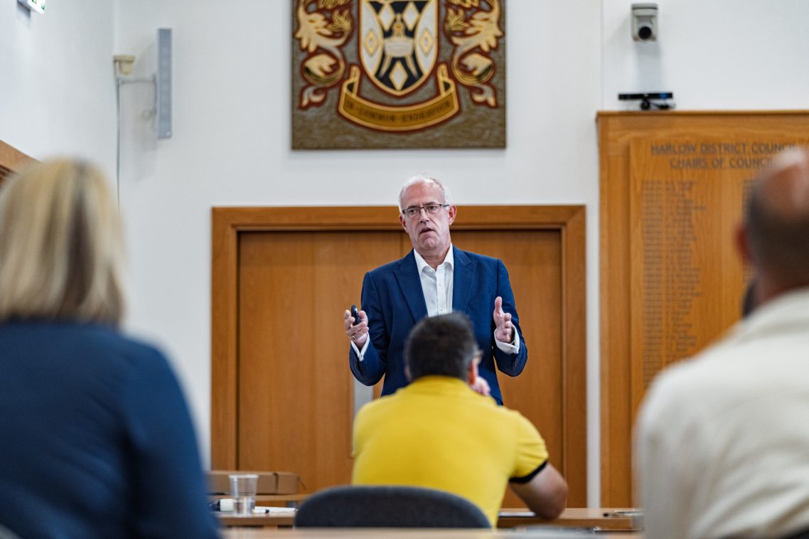 A speaker addresses an audience in a formal setting with a coat of arms on the wall.