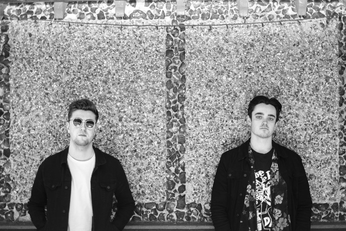 Two young men stand against a textured wall in a monochrome setting.