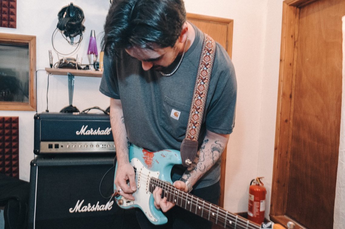 Man playing an electric guitar in a music studio with amplifiers in the background.