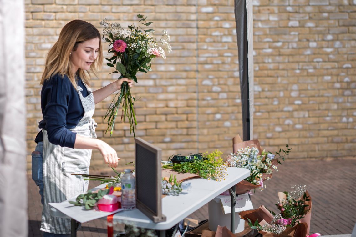 A woman arranging flowers at a table in a bright, outdoor setting.